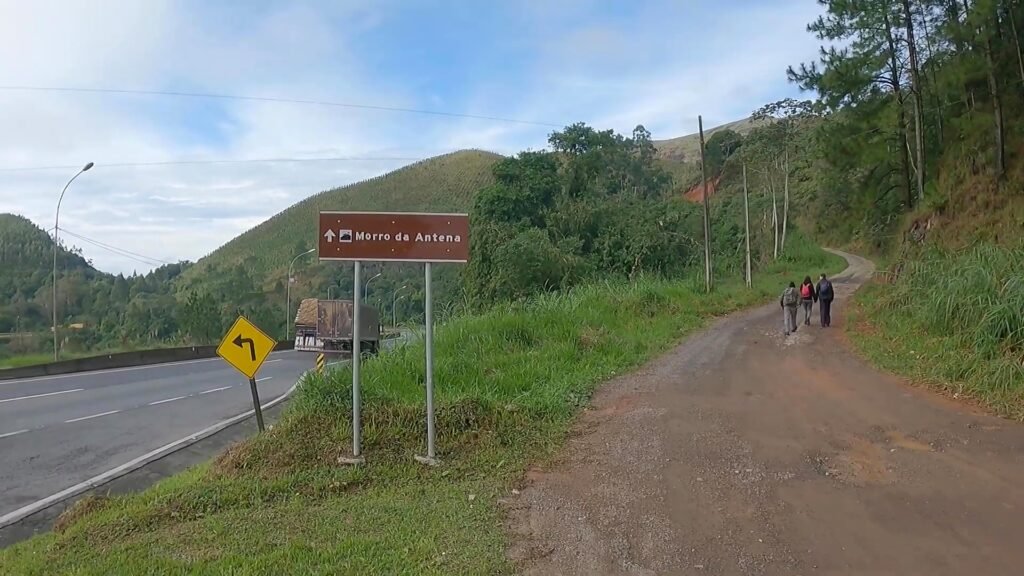Placa de sinalização "Morro da Antena" na beira da estrada, com trilha de terra subindo entre a mata e caminhantes ao fundo — local do encontro da Alcateia Lobo Guará em 29 de agosto.