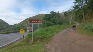 Placa de sinalização "Morro da Antena" na beira da estrada, com trilha de terra subindo entre a mata e caminhantes ao fundo — local do encontro da Alcateia Lobo Guará em 29 de agosto.